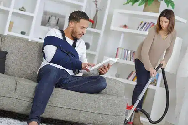A person with a arm sling, sits on a couch reading a book while another person vacuums around.